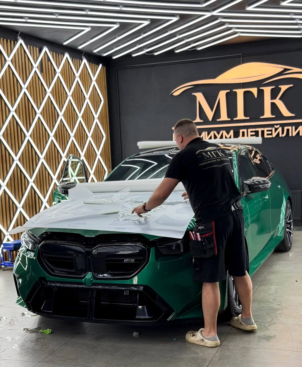 Мастер устанавливает прозрачную защитную плёнку на капот зелёного BMW