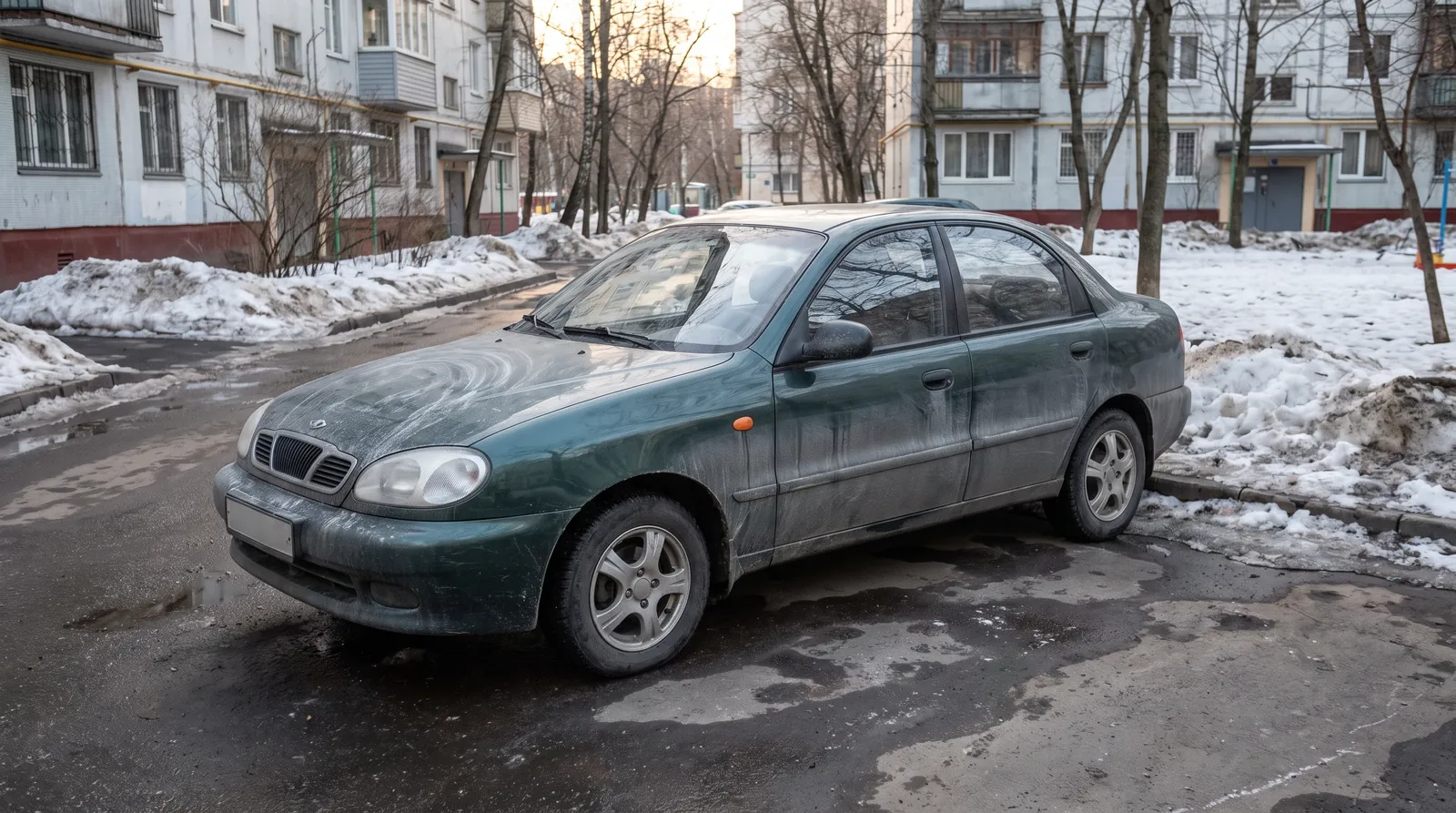 Тёмно-зелёный седан в московском дворе весной: кузов в белёсом солевом налёте на фоне тающих сугробов